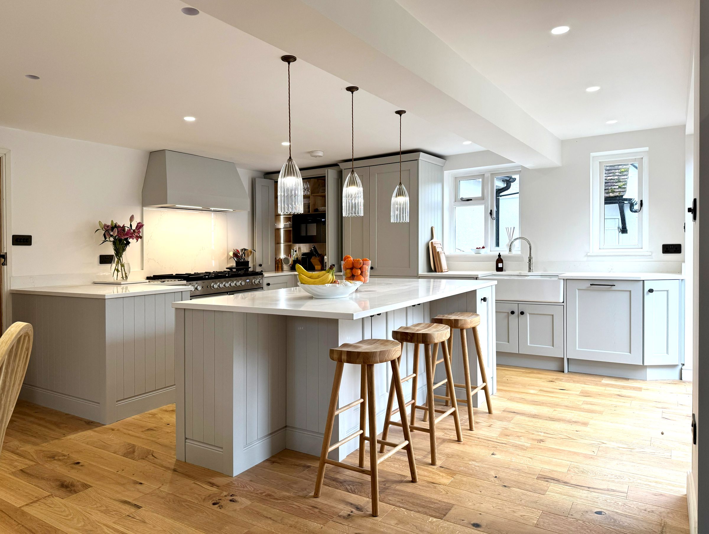 A pale painted shaker kitchen with an island, oak stools and glass pendants