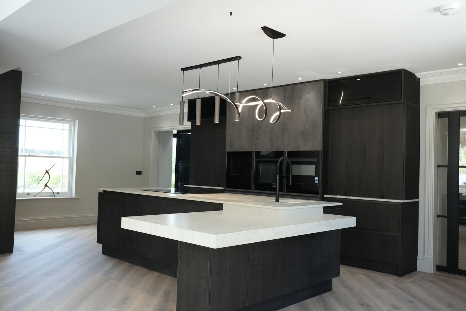 Wide view of an open-plan dark kitchen with a two-level white island and sculptural pendant light